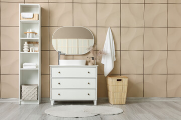 Chest of drawers, mirror and shelving unit near beige tile wall in stylish bathroom