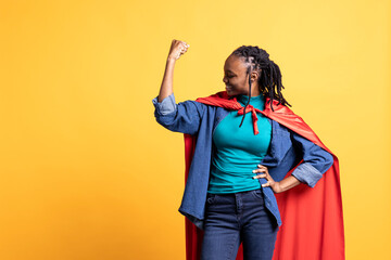 Happy BIPOC girl portraying superhero with red cape flexing muscles, isolated over studio...