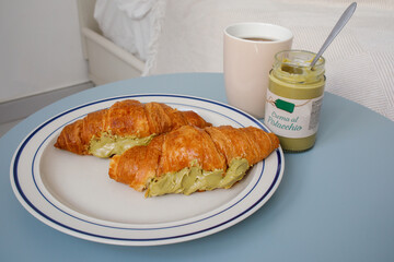 Two croissants filled with pistachio cream paste with black coffee on blue table near the bed