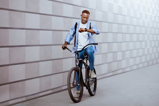 Happy African American guy with earphones listening to music and checking time on smartwatch during bike ride outside. Sporty black man riding modern bicycle on city street. Urban travel - Powered by Adobe