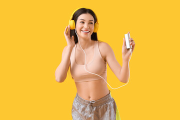 Sporty young woman with headphones and modern power bank on yellow background