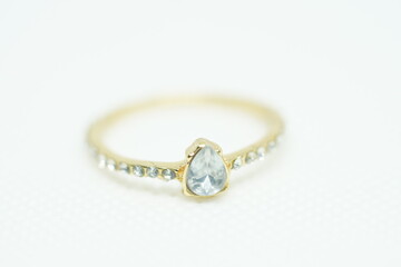 Close-up of a delicate gold ring with a teardrop-shaped clear gemstone and side stones, placed on a white textured background. Perfect for bridal, fashion, elegance, and luxury jewelry themes.