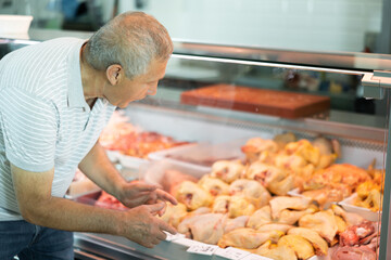Interested elderly man looking at large assortment of fresh raw meat products in glass refrigerated display case in butchery shop