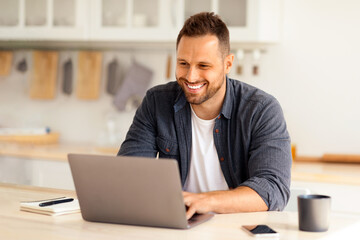 Cozy work at home Young happy man independent contractor working on laptop and smiling, enjoying remote work, sitting at home kitchen interior, free space