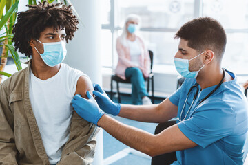 Obraz premium Man doctor putting medical plaster to African American guy patient's arm in clinic office after vaccination injection against covid-19 during pandemic. Vaccinating adults against corona virus concept