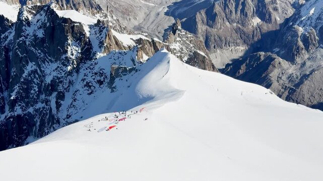 Paragliders with backpacks descend the steep snowy slopes of Aiguille du Midi, Chamonix, preparing for a parachute jump between the majestic peaks of the French Alps, with amazing view of Mont Blanc