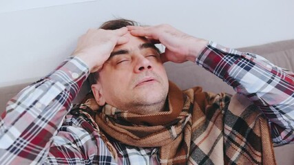 Man sitting on a couch with a pained expression, holding his forehead while wearing a scarf in a cozy indoor setting
