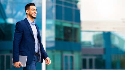 Happy young arab businessman walking from office, holding notepad, looking at copy space and...