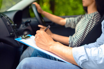Obraz premium Cropped of male instructor examinating female student, taking notes at test chart while sitting by young woman driving automobile, side view, copy space, closeup. Driving school concept