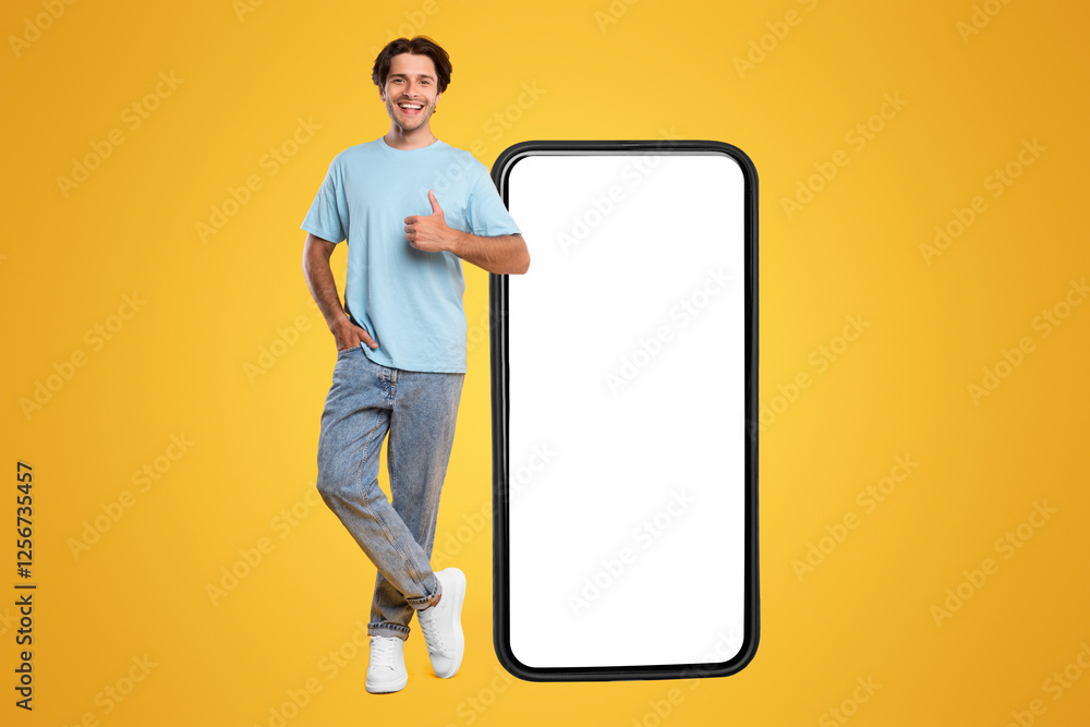 Poster happy man standing near big smartphone with empty white display gesturing thumb up sign, cheerful ca