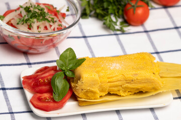 Humitas en hojas, plato típico Chileno en Verano, junto a ensalada de tomate decorada con hojas de albahaca, al fondo tomates maduros, perejil y una ensalada de tomate con cebolla