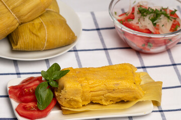 Humita en hojas, plato típico Chileno en Verano, junto a ensalada de tomate decorada con unas hojas de albahaca,  al fondo una ensalada de tomates con cebolla y perejil junto a un par de humitas