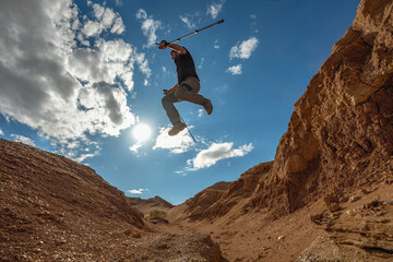 A man with trekking poles in hiking clothes jumps from one edge of a cliff to the other. A traveler's leap against the backdrop of a canyon and blue sky. Jump against the sky, view from the bottom.