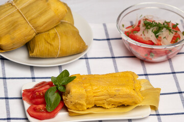 Humitas en hojas, plato típico Chileno en Verano, junto a ensalada de tomate y unas hojas de albahaca, al fondo una ensalada de tomates con cebolla y un plato con un par de humitas cerradas