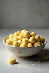 A bowl of creamy, pale yellow spheres, dusted with fine powder, rests on a light surface; a few more spheres lie scattered nearby, suggesting a sweet or savory treat.