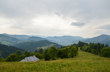Tranquil mountain scene with rolling hills, verdant trees, and charming rustic house under overcast...