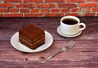 A piece of chocolate cake on a plate and a cup of hot black coffee on a wooden table, a dessert fork nearby.