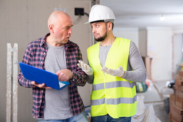 Puzzled apologetic young bearded construction worker talking with displeased man standing with...