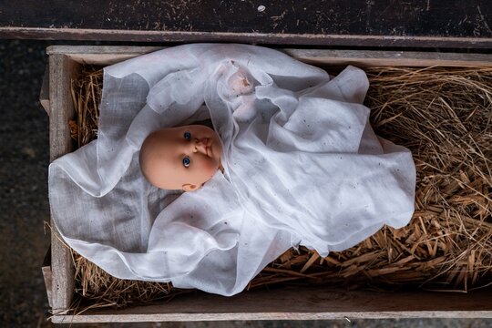 A baby doll, wrapped in a white cloth, rests in a wooden manger filled with hay. A still life nativity scene. Clevedon, Auckland, New Zealand