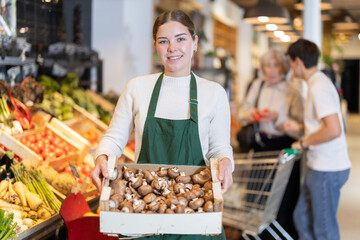 Girl employee in dark green apron stands in sales area with box of mushrooms. Employee carries box of mushrooms from storage area into sales area.