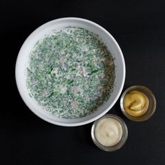 Classic okroshka on kefir in a white bowl with mustard and horseradish in glass sauce dishes, shot on a dark background