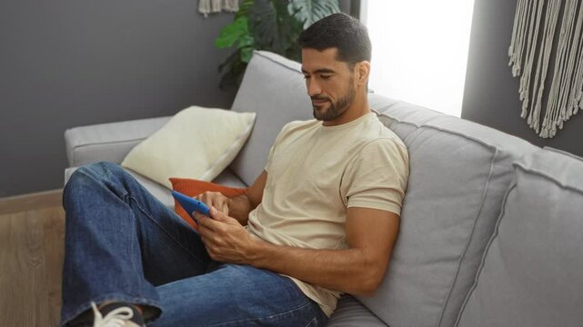 Young man with beard using tablet in living room shows a relaxed setting with modern decor and soft lighting, creating a cozy indoor atmosphere.
