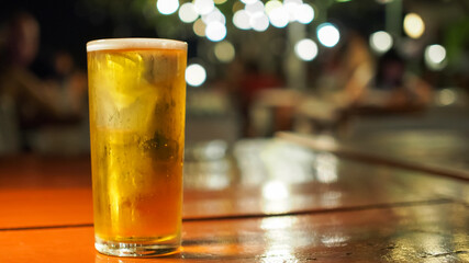 A crisp glass of golden beer placed on a glossy wooden table, with a blurred bokeh of warm lights and a cozy evening ambiance in the background