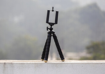Black Flexible mini octopus tripod with Bracket Holder Mount, placed on an outdoor wall with a blurred background. Compatible with smartphones and cameras.