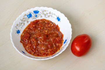 Tasty homemade Tomato Bhorta is served in a white bowl on a light wooden surface, with a whole red tomato resting beside it. Tomato bharta is a popular Bengali dish.