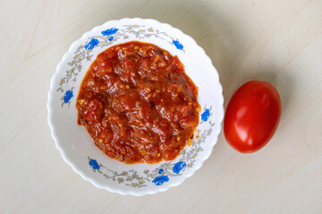 Homemade Tomato Bhorta, a popular Bengali dish, is presented in a white bowl on a light wooden surface, accompanied by a whole red tomato. Top view.