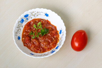 Delicious Tomato Bhorta is served in a white ceramic bowl on a light wooden surface, with a whole red tomato resting beside it. Tomato bharta is a popular Bengali dish.
