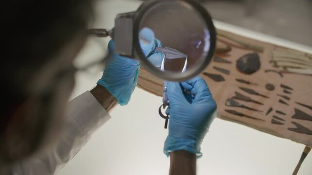 Archaeologist in gloves measuring ancient arrowhead with calipers under magnifying glass, working at laboratory table with excavation tools on it. Close-up view