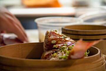 Close-up of raw fish topped with soy glaze, ginger, and scallions, placed inside a bamboo dim sum...