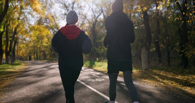 Rear view of a happy brunette guy in sportswear running along an alley in a sunny park in autumn with his girlfriend
