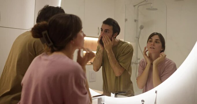 A girl and her boyfriend examine their face and preen themselves while standing in front of a mirror in a modern bathroom at home
