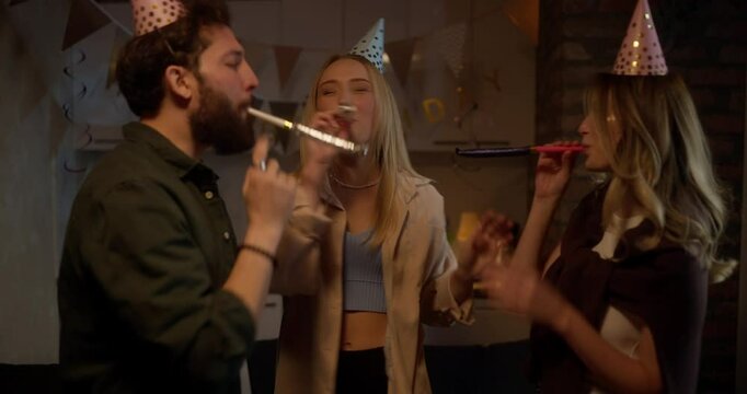 Cheerful girls together with their friend in a festive cap play and make noise with the help of Party Horn during a fun party at home in the evening