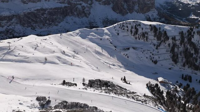 Alpine ski slopes wind down snow-covered mountain terrain in the Dolomites, with ski lifts and skiers dotting the pristine winter landscape against rugged mountain backdrop.
