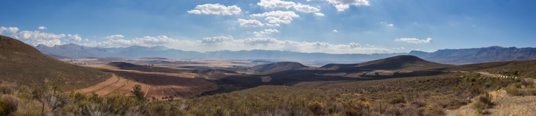 A panoramic view of the scenic Ceres Valley in South Africa, showcasing rolling hills, fertile farmlands, and distant mountain ranges under a bright blue sky with scattered clouds.
