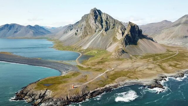 Breathtaking aerial view capturing the dramatic Hvalnes landscape in Iceland., featuring a vibrant orange lighthouse on a rocky coast a tranquil nature reserve and majestic mountains under a clear sky