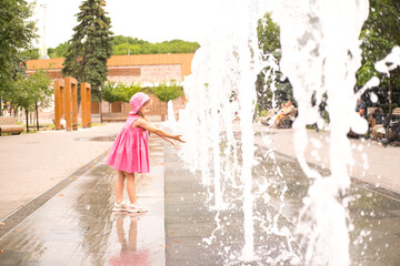 kid girl in pink dress plays next to fountain on summer day