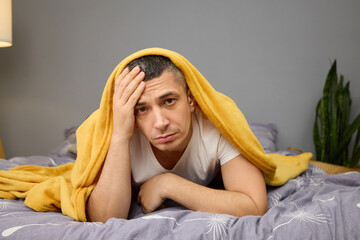 A man lies on a bed with a yellow blanket draped over him, looking tired and stressed. The room is...