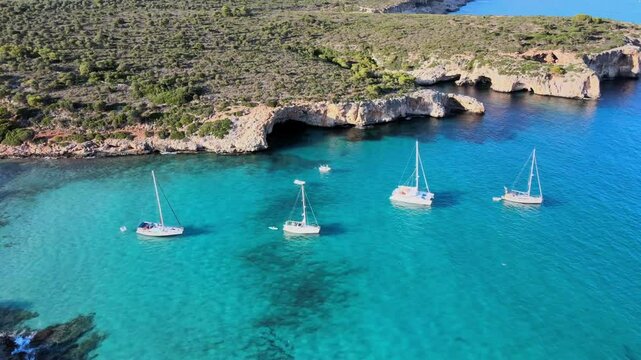 Experience the stunning beauty of Cala Varques, located near Porto Cristo in Majorca. Sailboats float serenely in the vibrant turquoise waters, surrounded by lush greenery and rocky cliffs.