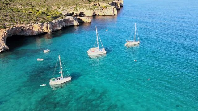 Clear turquoise waters surround sailboats anchored near the stunning cliffs of Cala Varques. This serene location in Majorca offers breathtaking views and tranquil vibes perfect for relaxation.