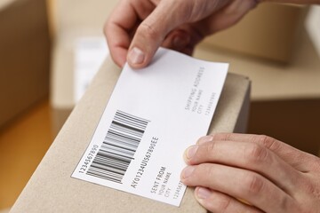 Obraz premium Man sticking shipping label with barcode on parcel indoors, closeup