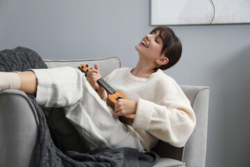 Happy woman playing ukulele in armchair at home