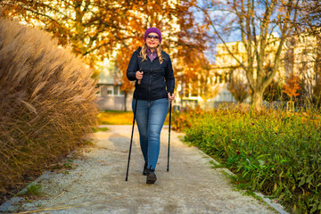 Beautiful middle aged woman practicing nordic walking in city park on autumn day. Front view....
