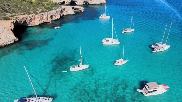Experience the tranquil beauty of Cala Varques, Majorca, featuring numerous sailing boats anchored in crystal-clear waters. Enjoy the surrounding cliffs and lush greenery under a clear blue sky.
