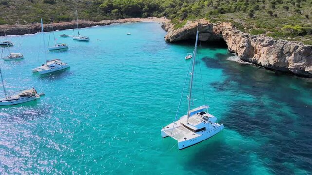 Experience the tranquil beauty of Cala Varques in Mallorca. Sailboats glide over crystal-clear turquoise waters surrounded by stunning cliffside landscapes. Perfect for boat lovers.