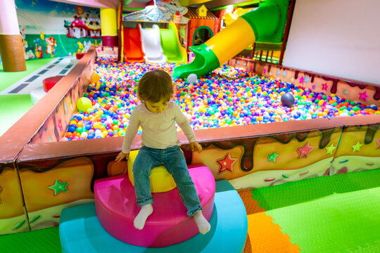 A child sitting on a soft play structure near a colorful ball pit in a vibrant indoor playground.