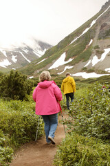 Senior couple in bright tourist jackets is hiking in lush greenery near spectacular mountains with snow. Trekking in wild places. Happy active retirement concept. Old people healthy habits. 